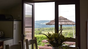 A dining area with a table, chairs, and a vase of flowers. A large window offers a view of a thatched umbrella and a scenic landscape. - Rhiwddu Barns - Brecon Beacons National Park (Llangadog)