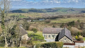 A stone house with a sloped roof, surrounded by a fenced yard and a view of rolling hills and a lake in the distance. - Rhiwddu Barns - Brecon Beacons National Park (Llangadog)