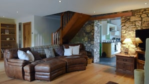 A living room with a brown leather sectional sofa, a wooden staircase, a stone wall, and a kitchen in the background. - Rhiwddu Barns - Brecon Beacons National Park (Llangadog)