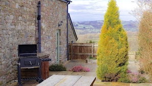 A stone building with a barbecue grill, a wooden fence, and a garden with a tall coniferous tree. - Rhiwddu Barns - Brecon Beacons National Park (Llangadog)