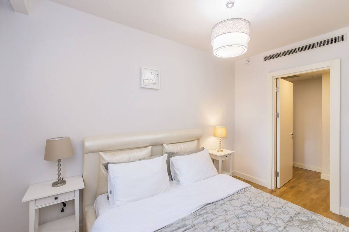 Ein Schlafzimmer mit einem Bett, zwei Nachttischen, einer Lampe, einer Deckenleuchte und einer Tür zu einem anderen Raum.