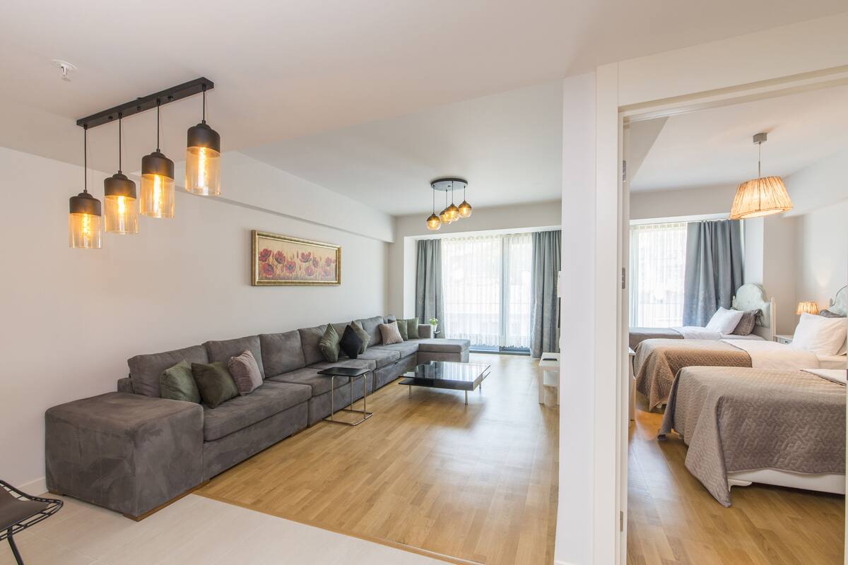 Ein modernes Wohnzimmer mit einer grauen Eckcouch, einem schwarzen Couchtisch und Holzboden. Es gibt eine Essecke mit weißem Tisch und Stühlen. Der Raum hat ein großes Fenster mit Vorhängen und einer Deckenleuchte.