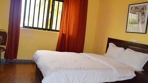 A neatly made bed with white linens in a room with yellow walls and a window with red curtains. - Hôtel Saint-Jean (Cotonou)