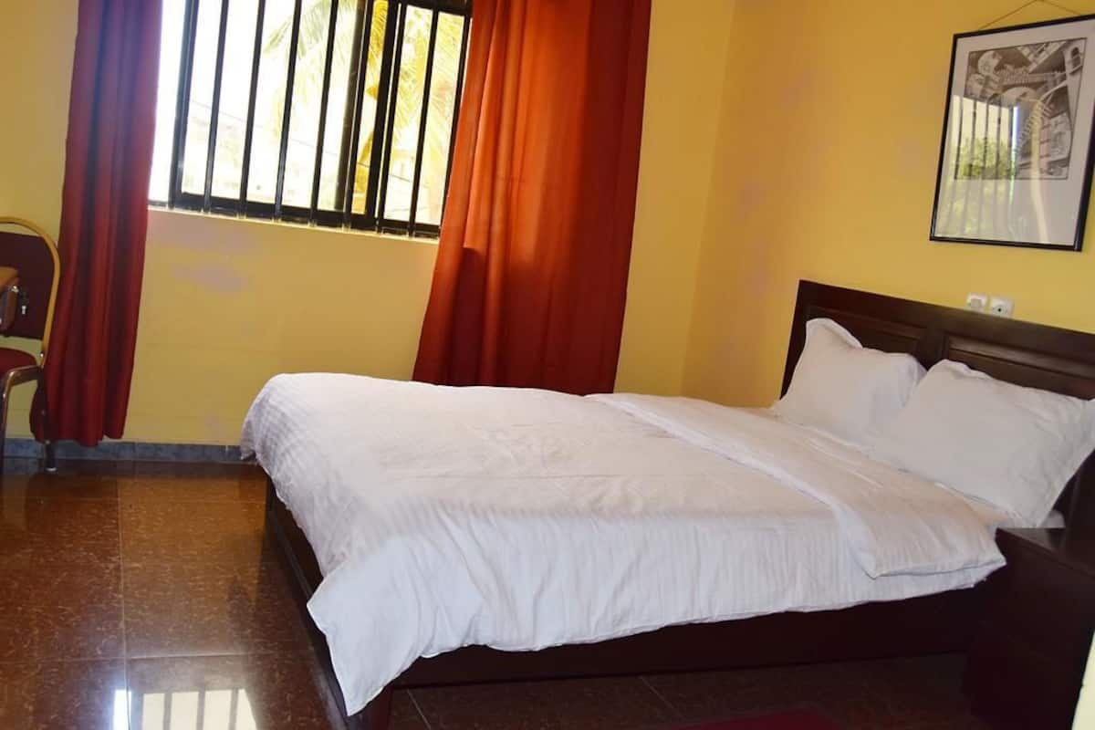 A neatly made bed with white linens in a room with yellow walls and a window with red curtains.