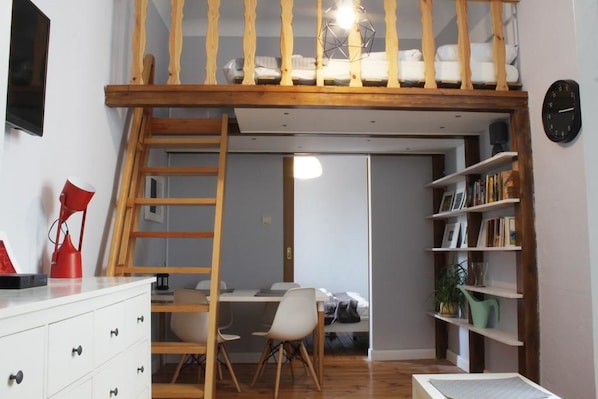 A loft bedroom with a wooden ladder, a desk, a chair, and a bookshelf.