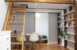 A room with a wooden ladder, a desk with chairs, a bookshelf, and a hanging light fixture.