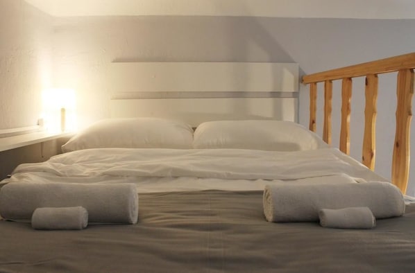 A neatly made bed with pillows and rolled towels, a wooden headboard, and a bedside lamp.