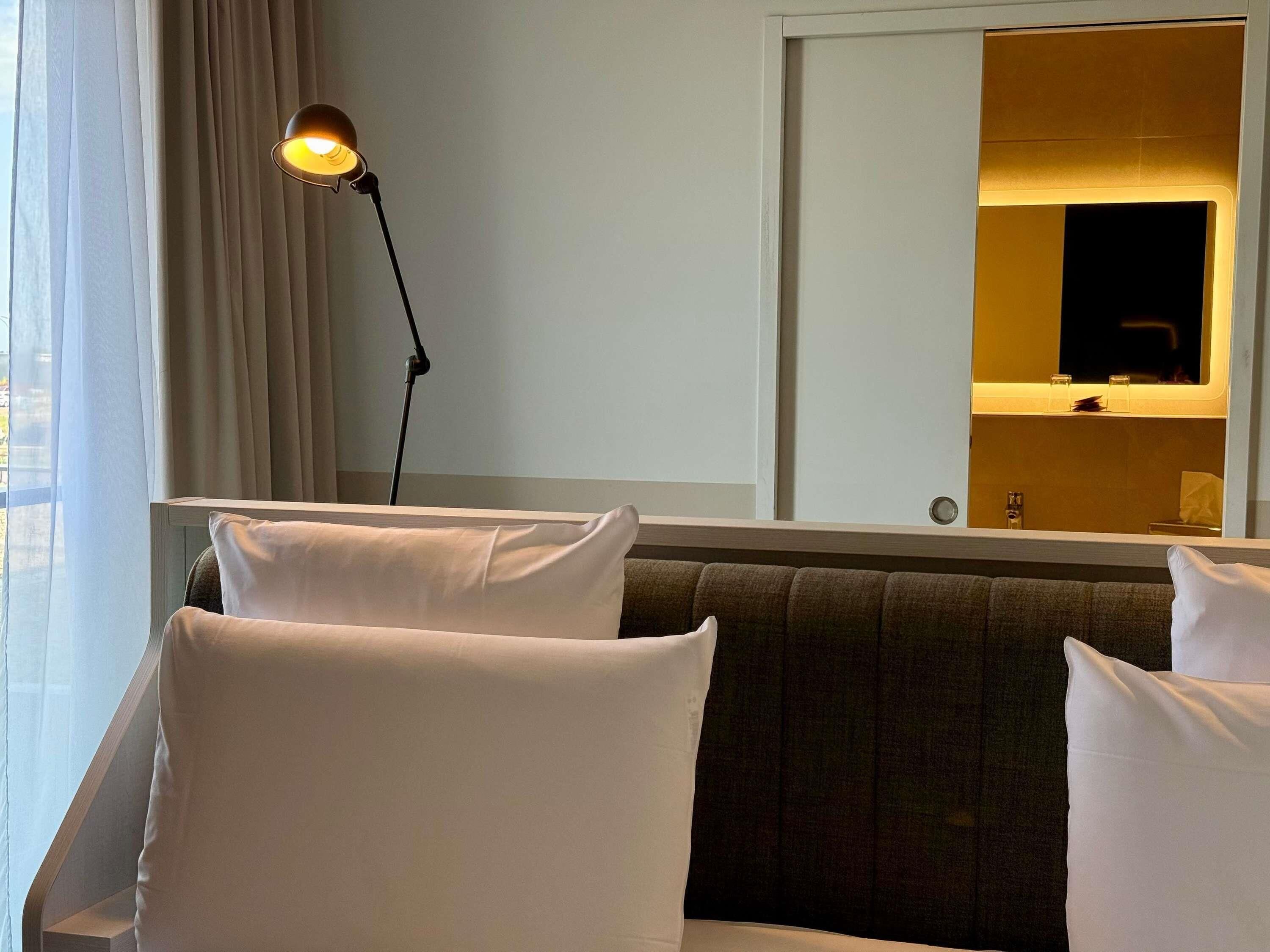 Habitación de hotel con sofá, lámpara de pie, espejo y ventana con cortinas.