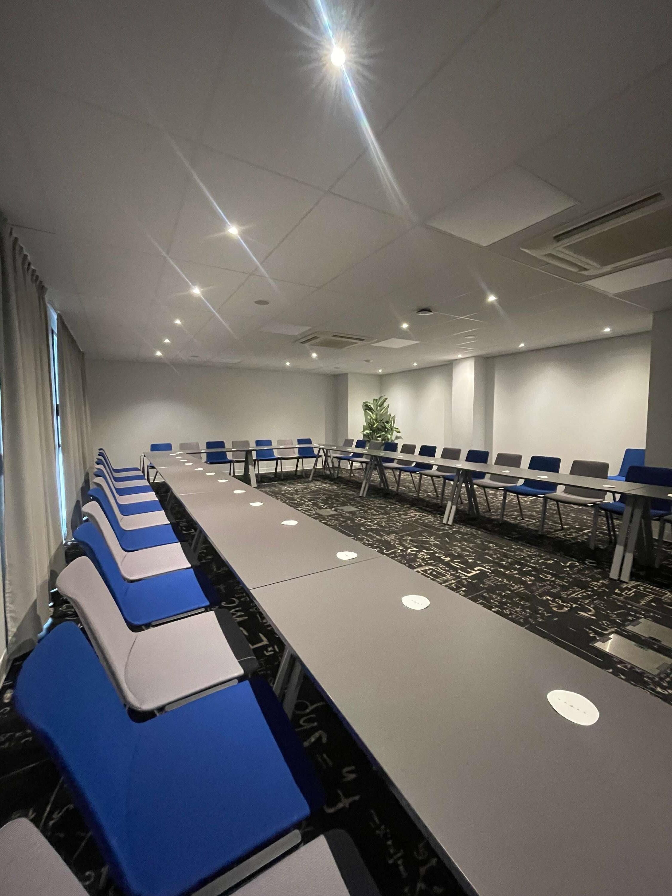 A conference room with rows of tables and chairs, a patterned carpet, and a ceiling with recessed lighting.