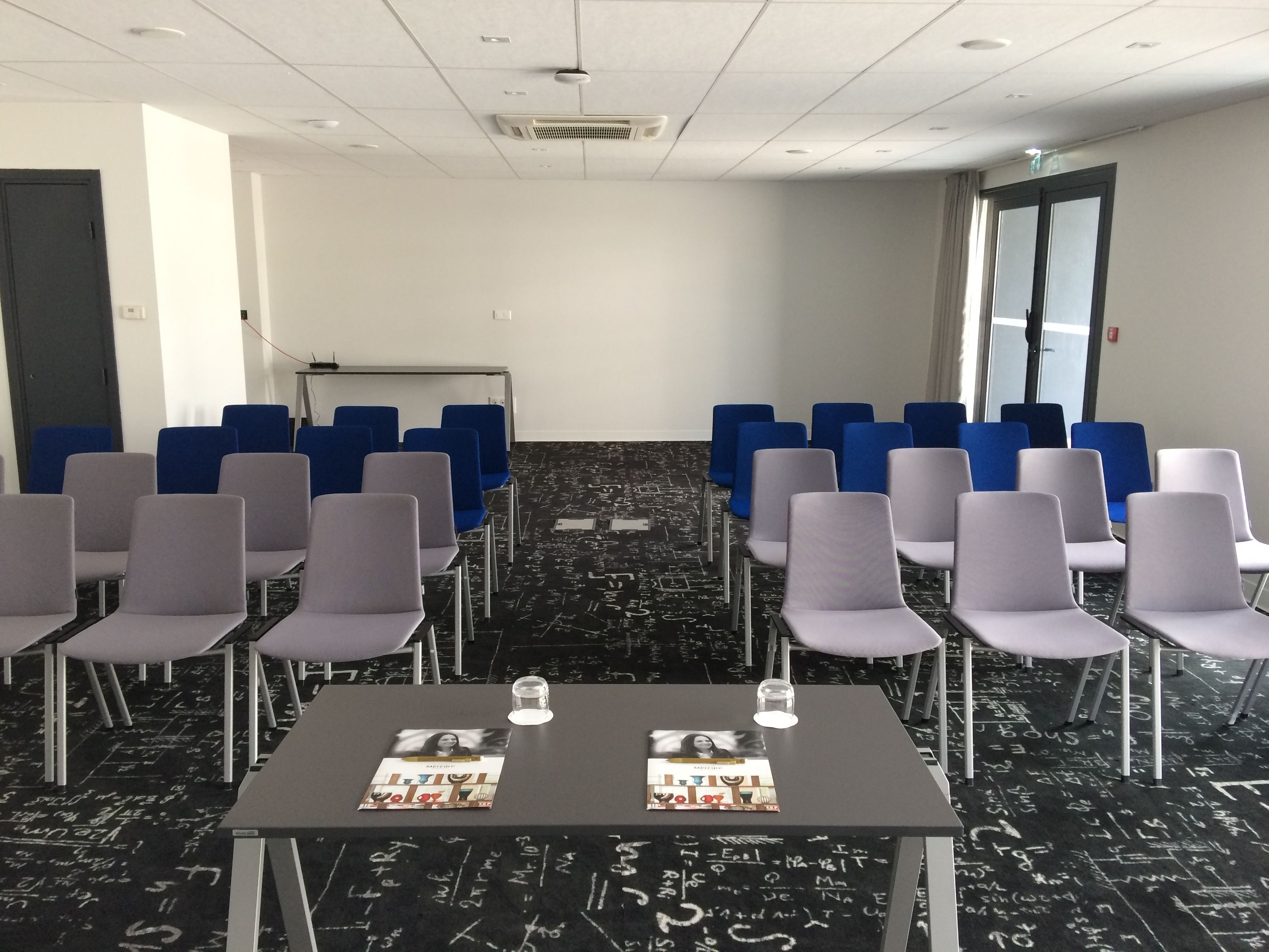 Un salón de conferencias con filas de sillas, una mesa con folletos y un piso negro con marcas similares a tiza blanca.