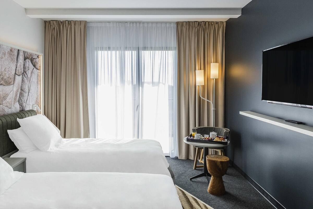Habitación de hotel con dos camas, un televisor, una mesita de noche con una bandeja de desayuno y un ventanal con cortinas.