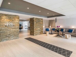 A modern hotel lobby with a stone accent wall, a seating area with sofas and a coffee table, and a visible kitchen in the background. - Mercure Hotel & Spa Bastia Biguglia (Biguglia)