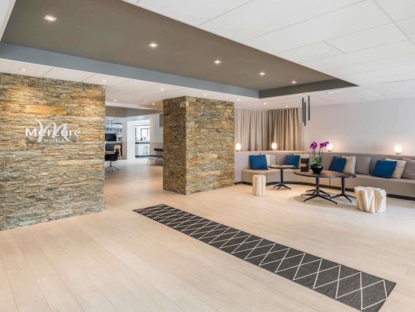 A modern hotel lobby with a stone accent wall, a seating area with sofas and a coffee table, and a visible kitchen in the background.