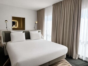 Ein modernes Hotelzimmer mit einem großen Bett, zwei Nachttischlampen, einem Spiegel und Blick auf die Stadt durch transparente Vorhänge.