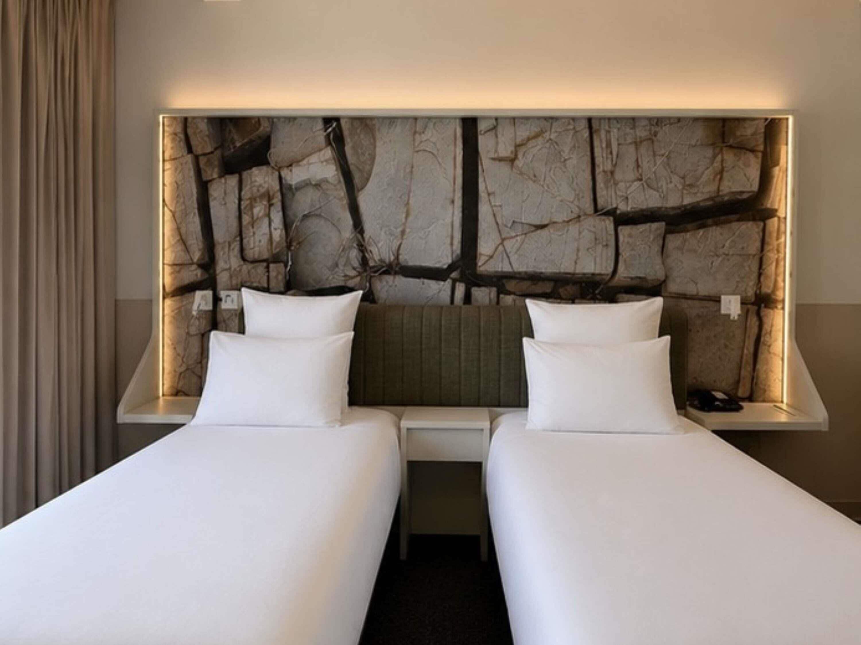 Une chambre d’hôtel avec deux lits, une tête de lit moderne et une table de chevet avec une lampe.