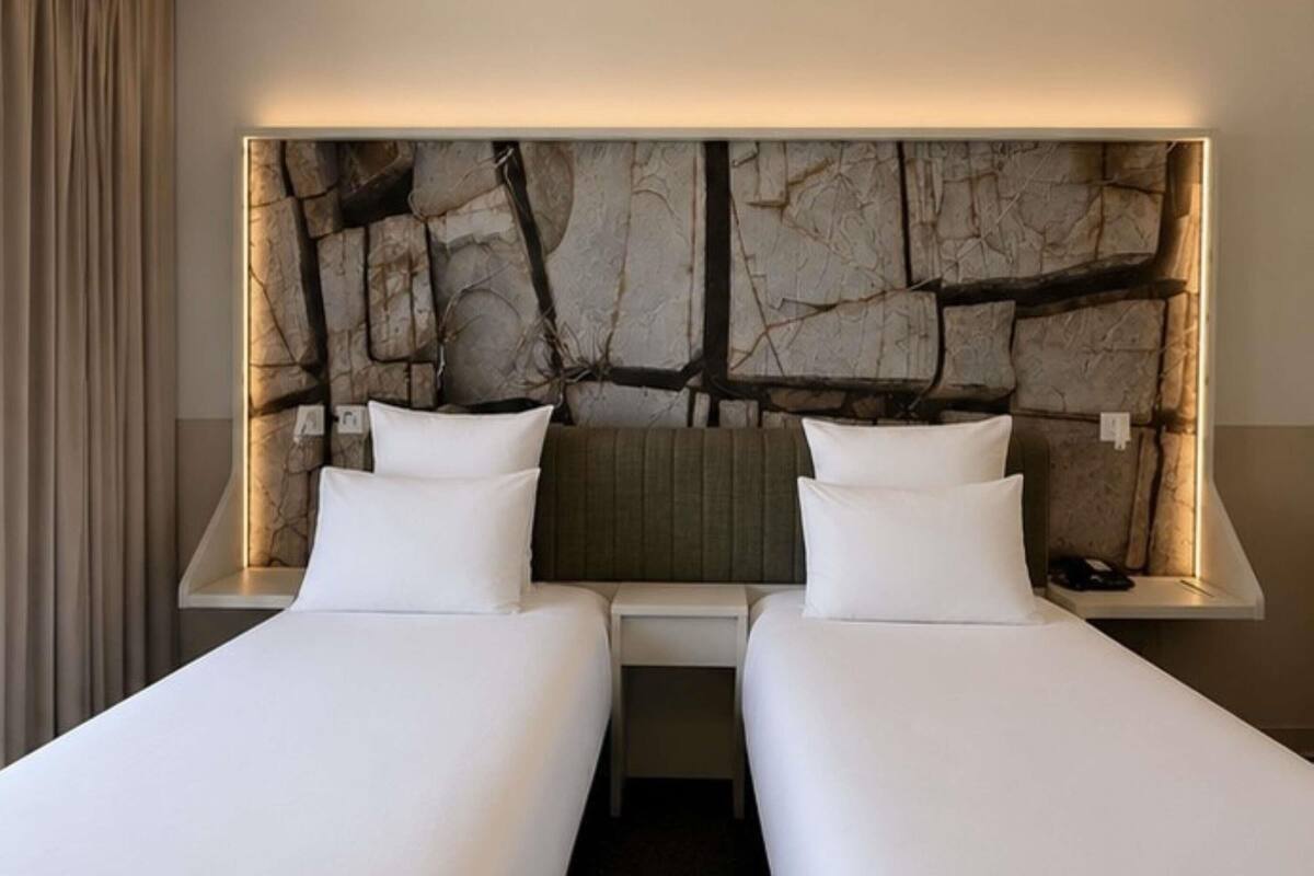 Una habitación de hotel con dos camas, una cabecera moderna y una mesita de noche con lámpara.