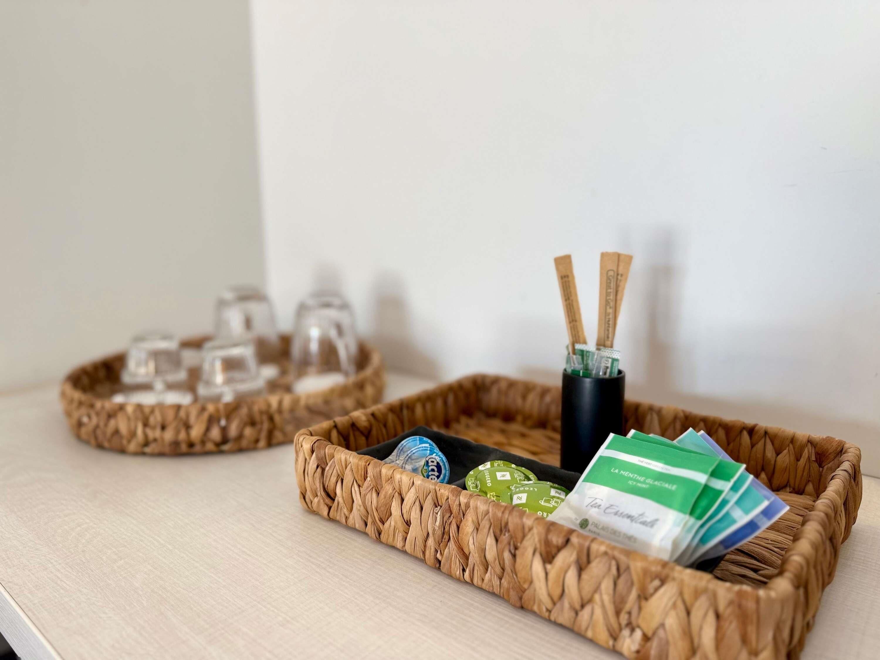 Deux paniers tressés sont posés sur une table : l’un contient des sachets de thé et un récipient noir avec des diffuseurs à roseaux, et l’autre contient des verres transparents et une bouteille.