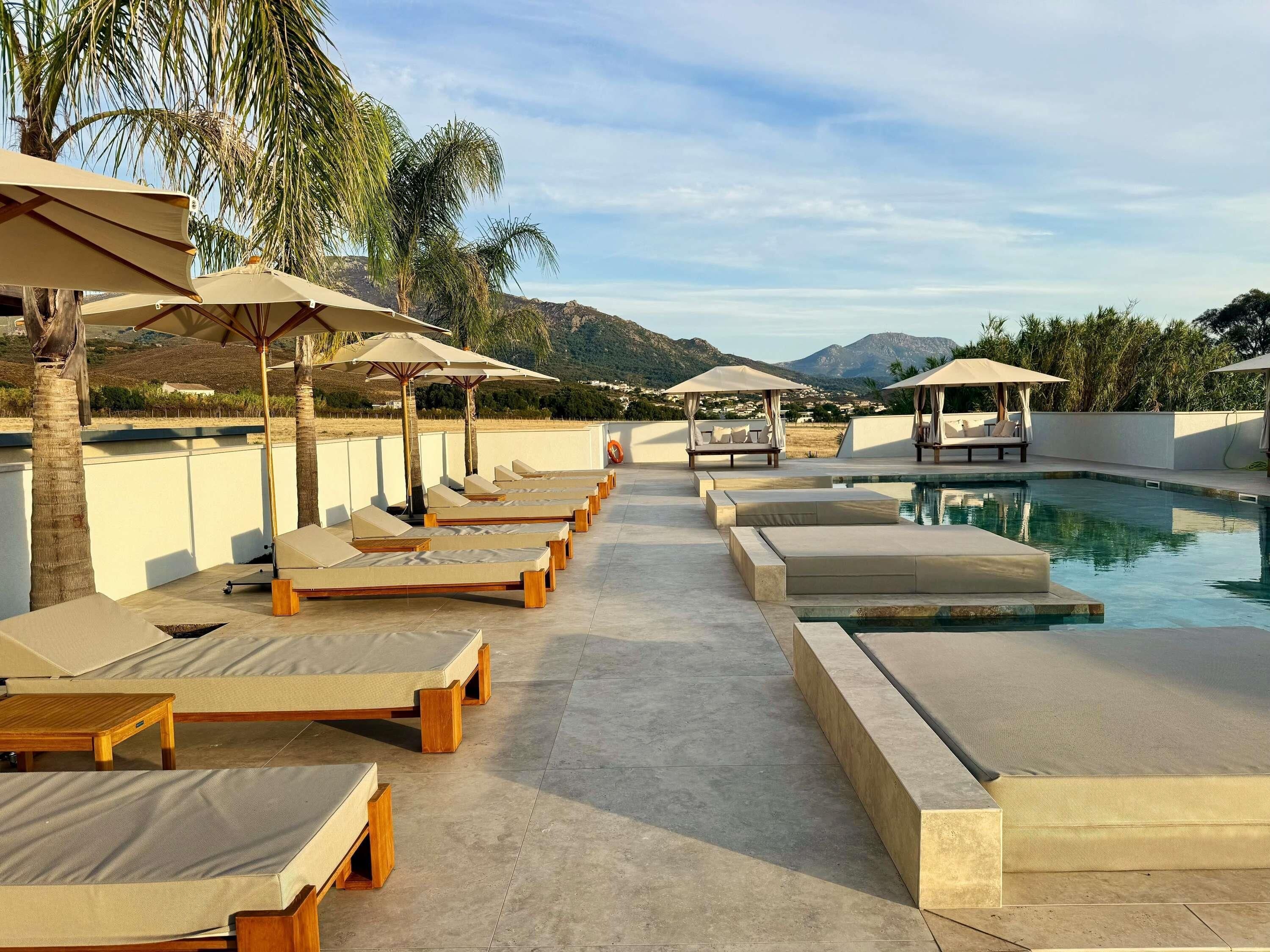 Zona de piscina con sillones de descanso y sombrillas, cielo despejado y montañas al fondo.