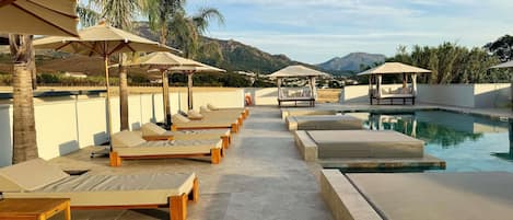 A poolside area with lounge chairs and umbrellas, a clear sky, and a mountainous backdrop.