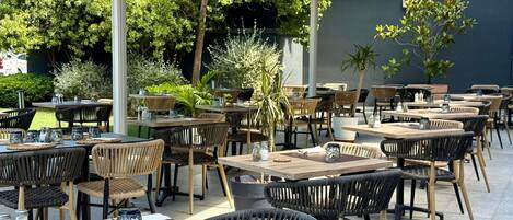 An outdoor dining area with multiple tables and chairs, surrounded by greenery and a building wall.