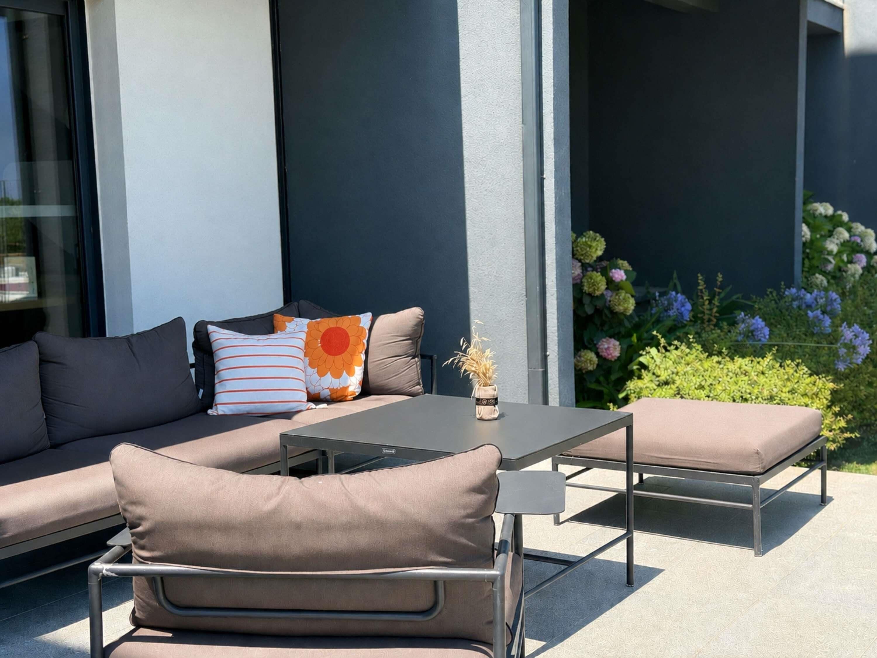 Un espace extérieur aménagé avec un canapé, des fauteuils et une table, entouré d’un jardin fleuri.