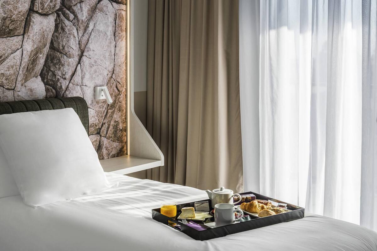 Una cama bien tendida con una bandeja de desayuno, que incluye croissants y jugo, en una habitación de hotel con pared de piedra y cortinas.