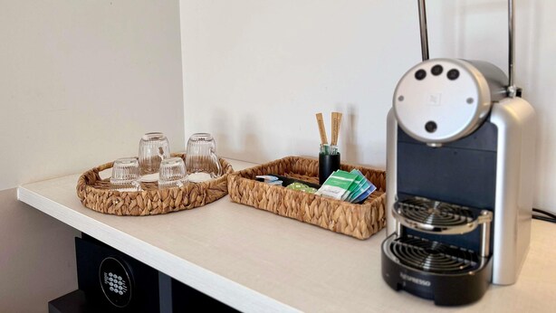 Eine Kaffeemaschine, zwei geflochtene Tabletts mit Gläsern und einer Flasche, sowie ein Korb mit Karten auf einem Tisch.