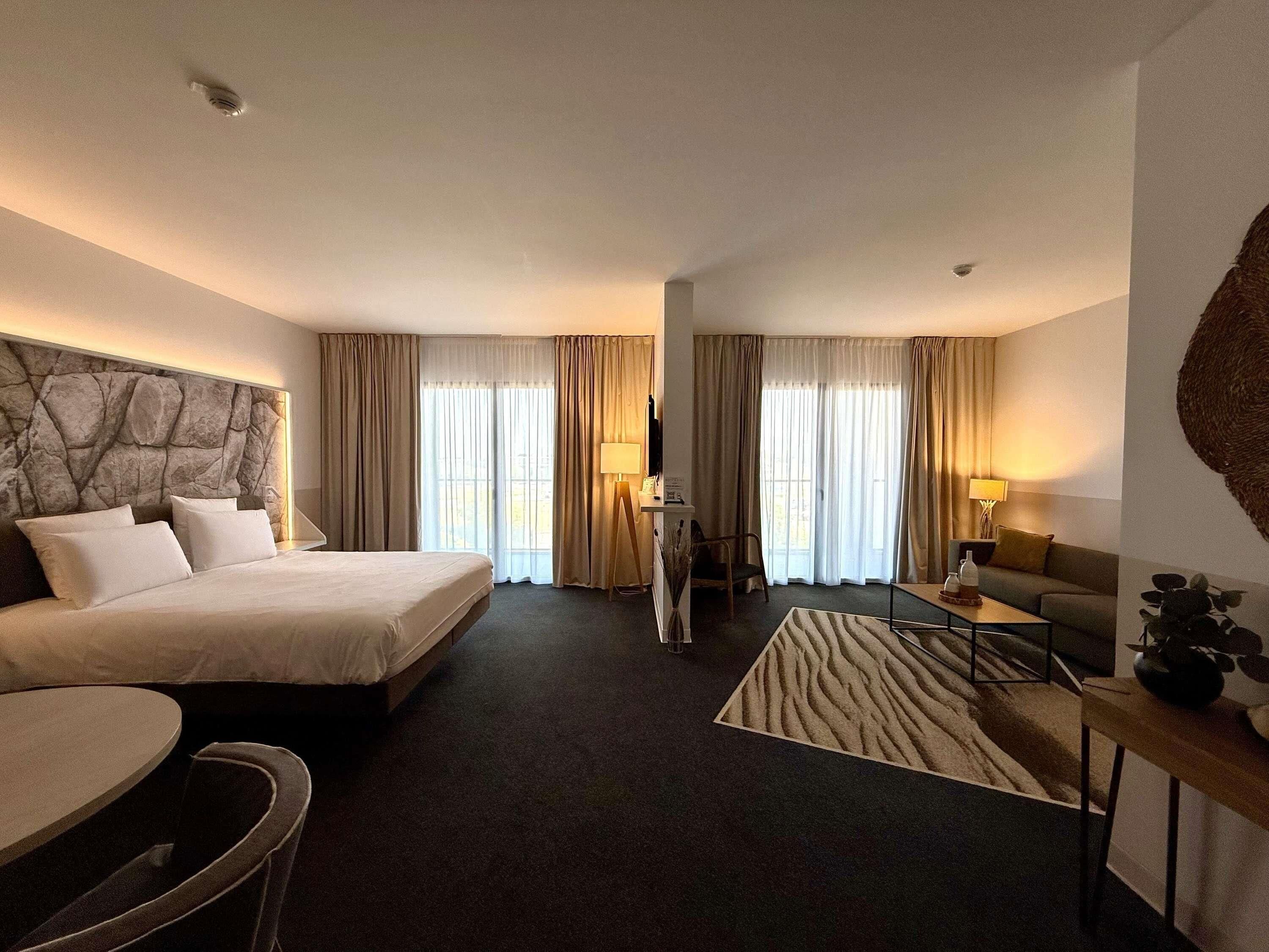 Ett hotellrum med en säng, ett skrivbord, en stol, en soffa och ett stort fönster med gardiner.