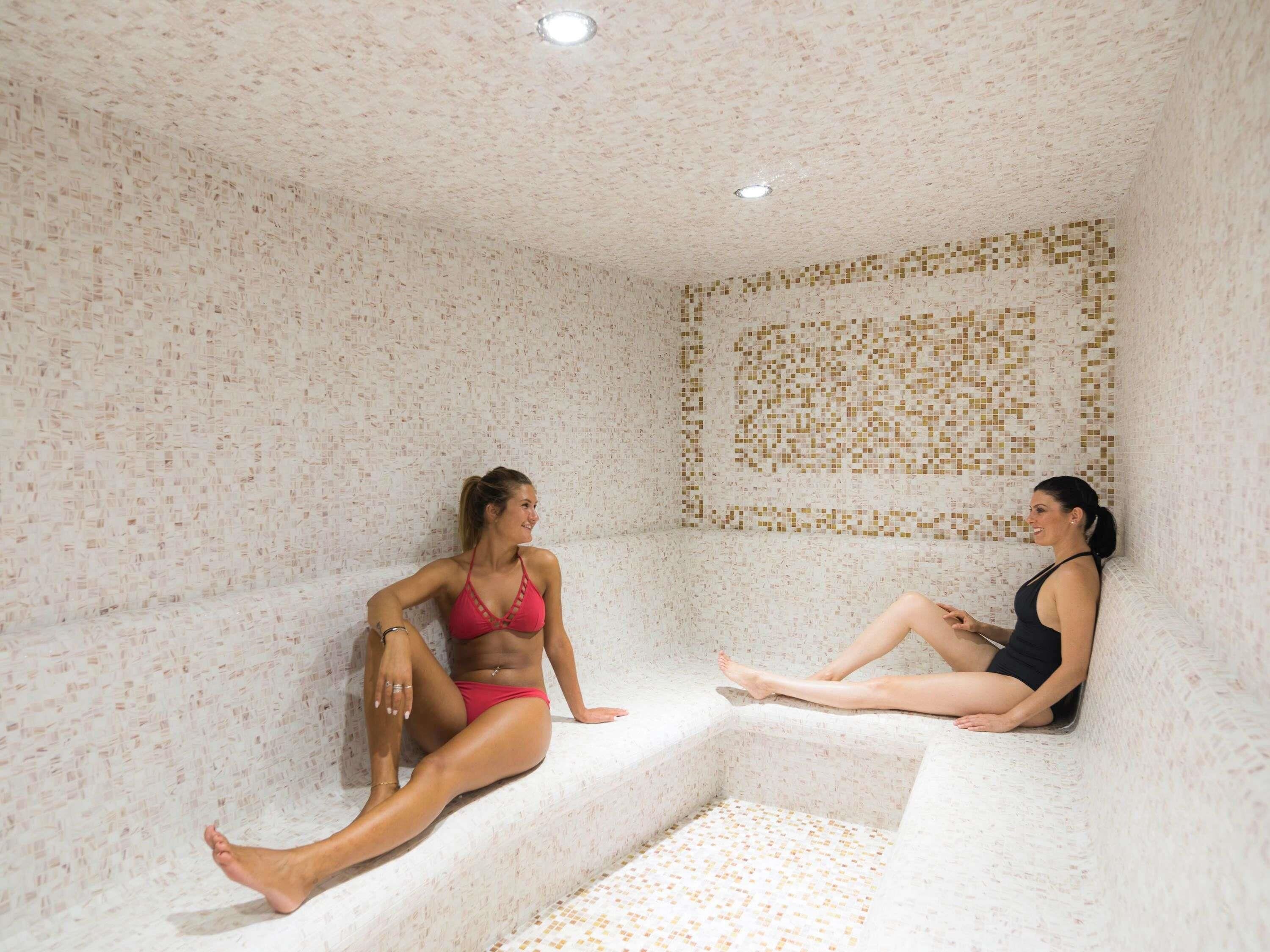 Dos mujeres sentadas en un espacio tipo spa con paredes de azulejos y un escalón que baja a un nivel inferior.