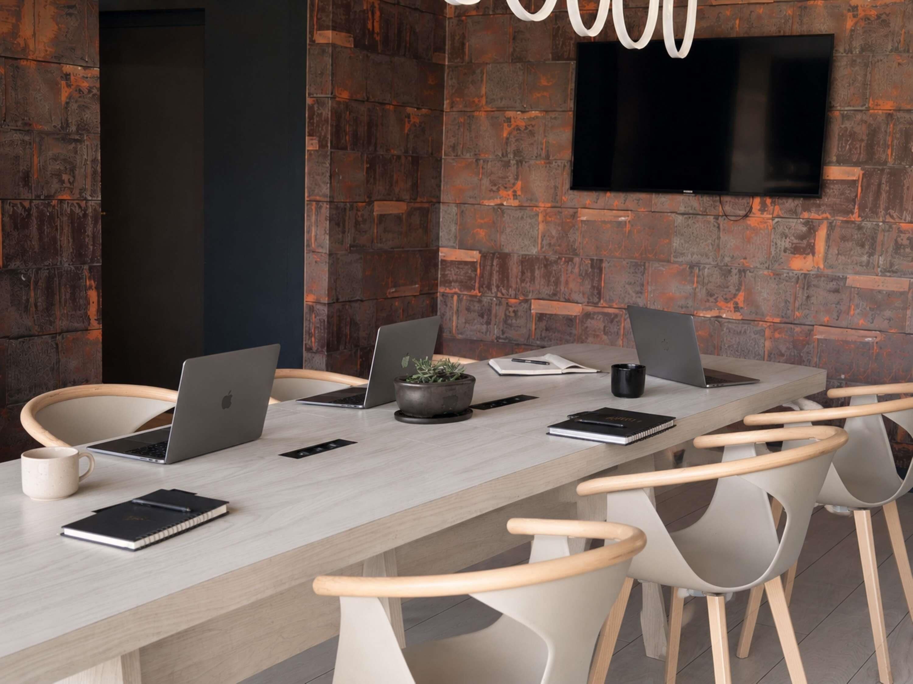 A conference room with a long table, laptops, chairs, a potted plant, and a flat-screen TV mounted on the wall.