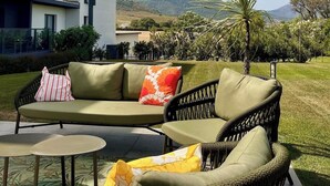Ein moderner Außenbereich mit einer Sitzecke, bestehend aus einer Couch und zwei Sesseln, einem kleinen Tisch sowie Blick auf ein Gebäude und Hügel im Hintergrund.