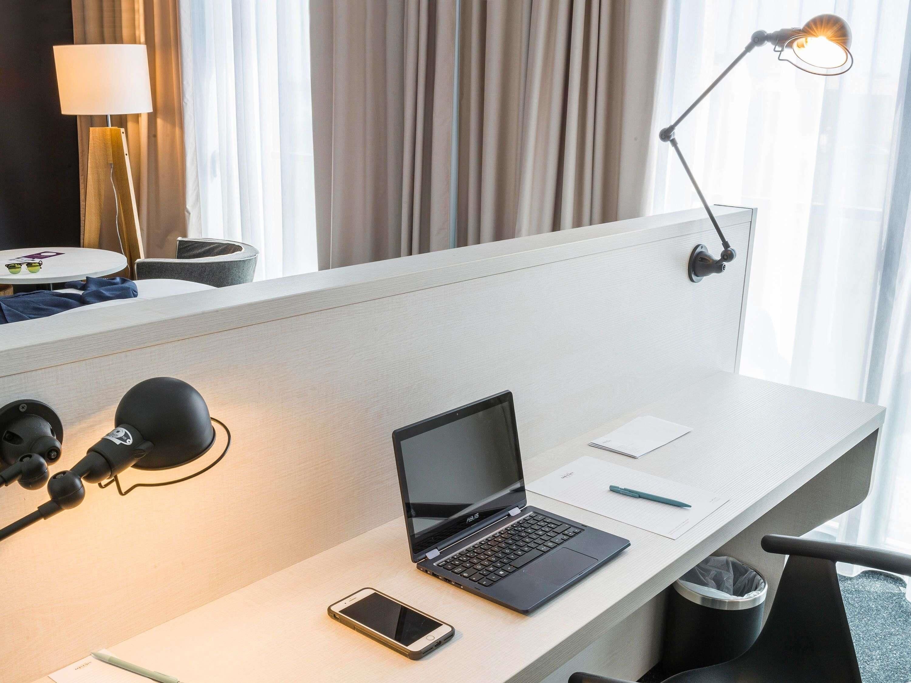 Un espacio de trabajo en un hotel con una computadora portátil, un teléfono inteligente, una lámpara de escritorio y un cesto de basura.