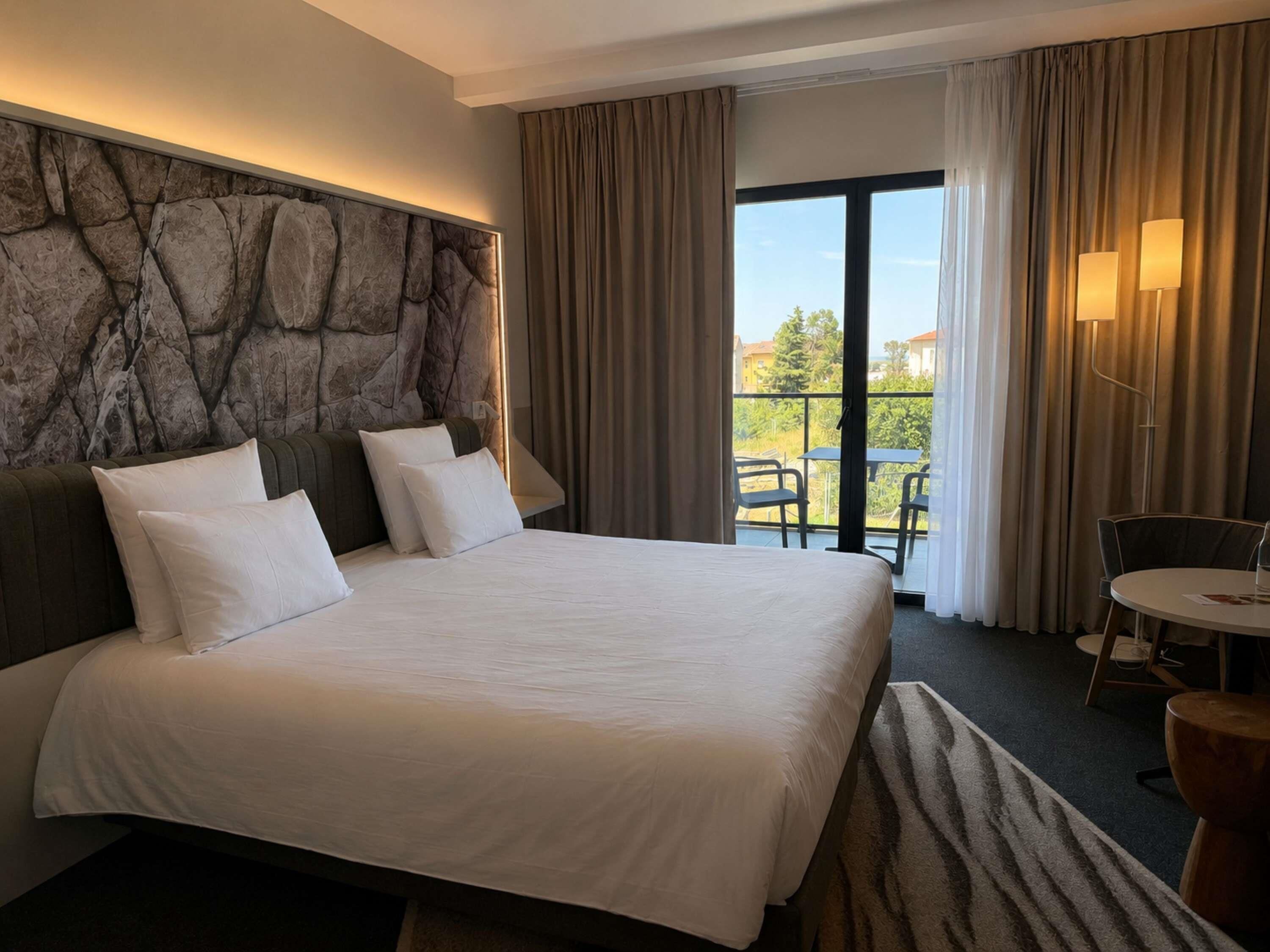 Habitación de hotel con una cama grande, una pared de acento de piedra, un balcón con mobiliario exterior y vistas al exterior.