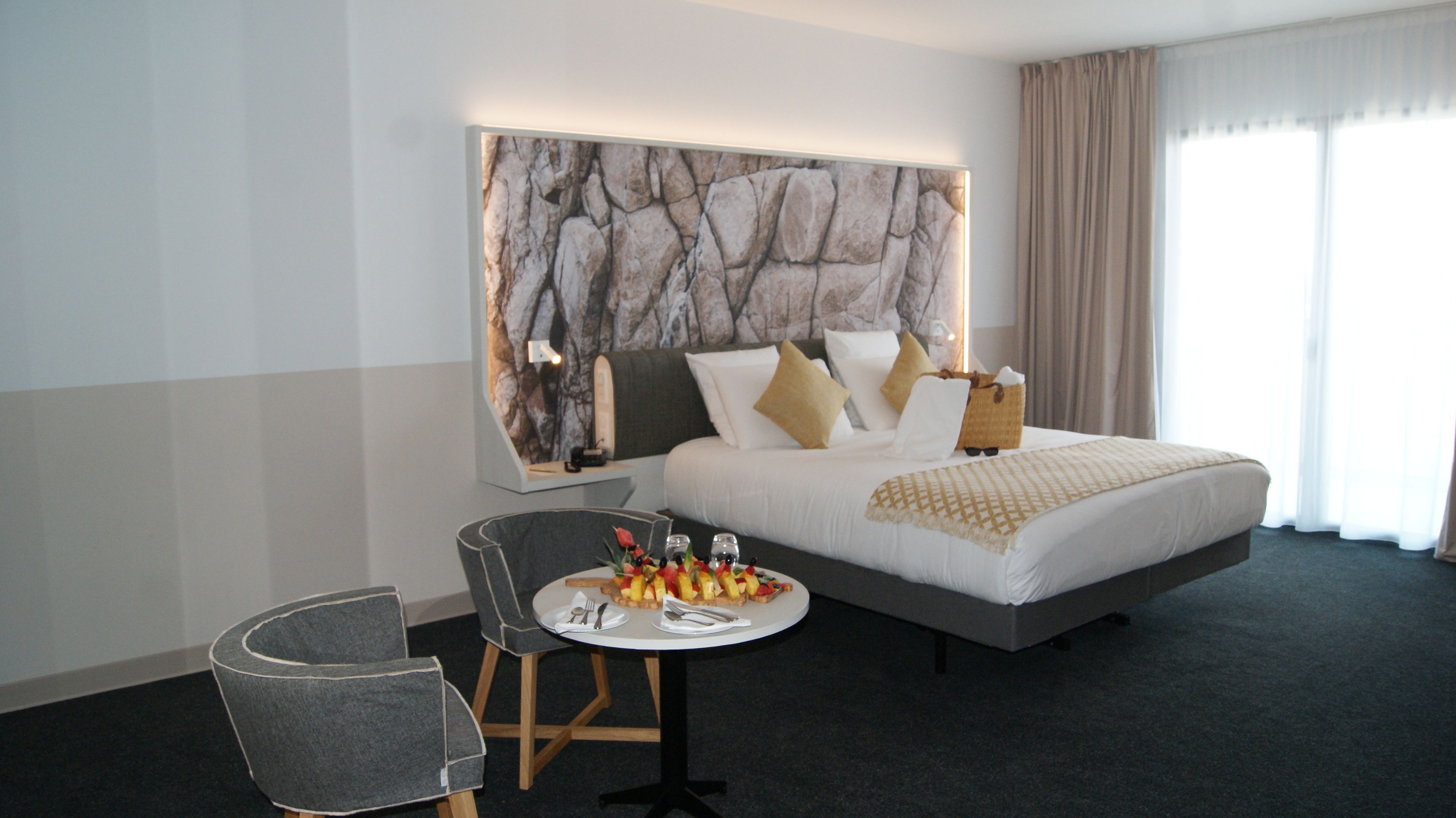 Habitación de hotel con una cama grande, una pequeña mesa redonda con desayuno, un sillón gris y una pared con diseño de mármol.