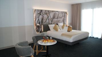 A hotel room with a large bed, a small round table with breakfast, a gray armchair, and a marble-patterned wall.