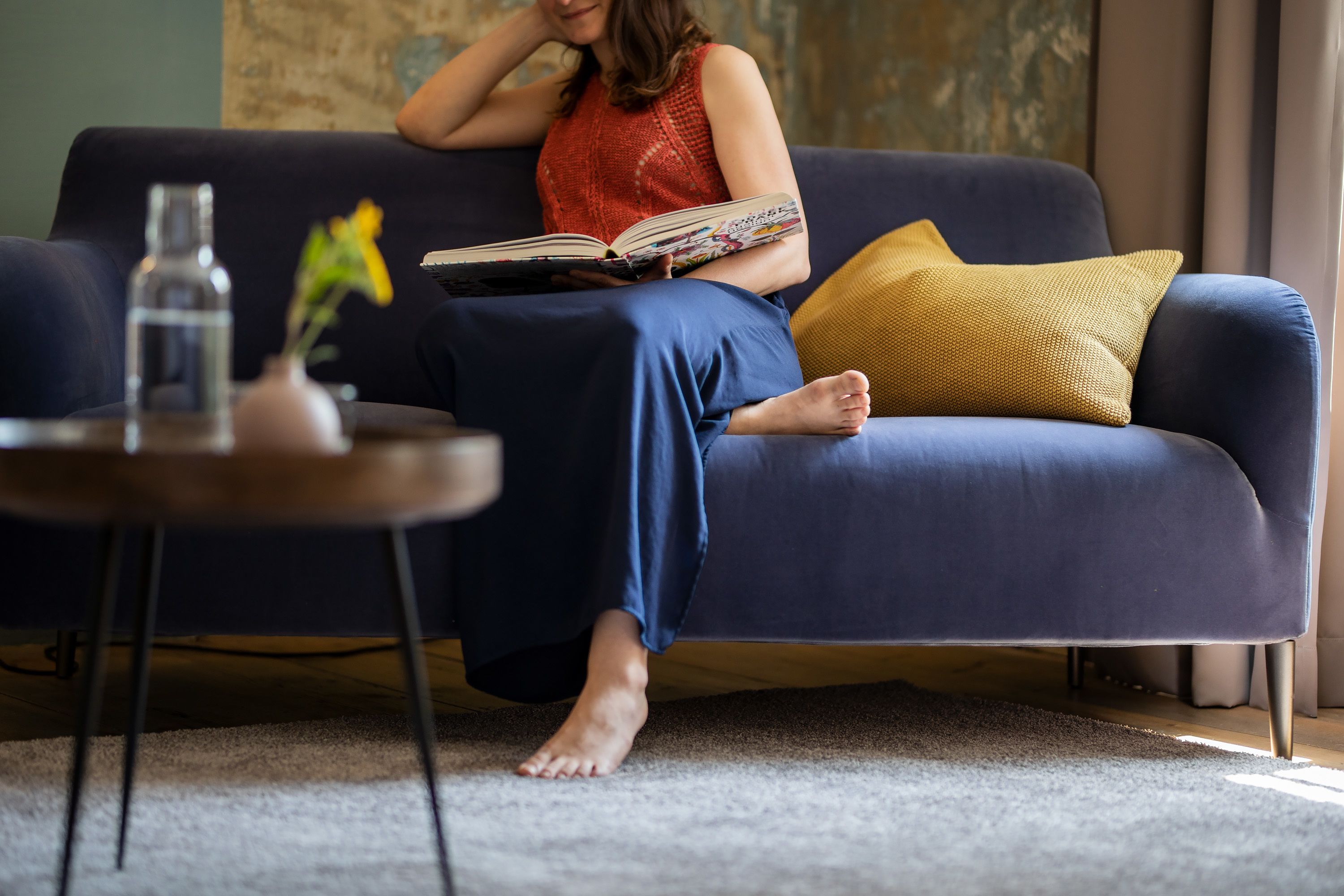 A person sitting on a blue sofa with a book, a yellow pillow, and a small table with a vase and a bottle.