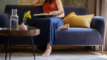 A person sitting on a blue sofa with a book, a yellow pillow, and a small table with a vase and a bottle.