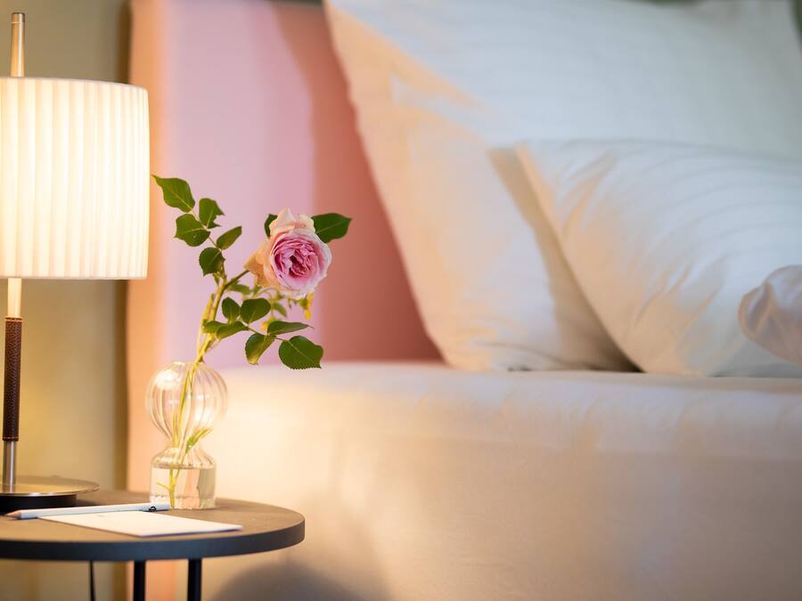 Una cama bien tendida con sábanas blancas, una mesita de noche con una lámpara y un florero con una sola rosa, y una almohada rosa al fondo.