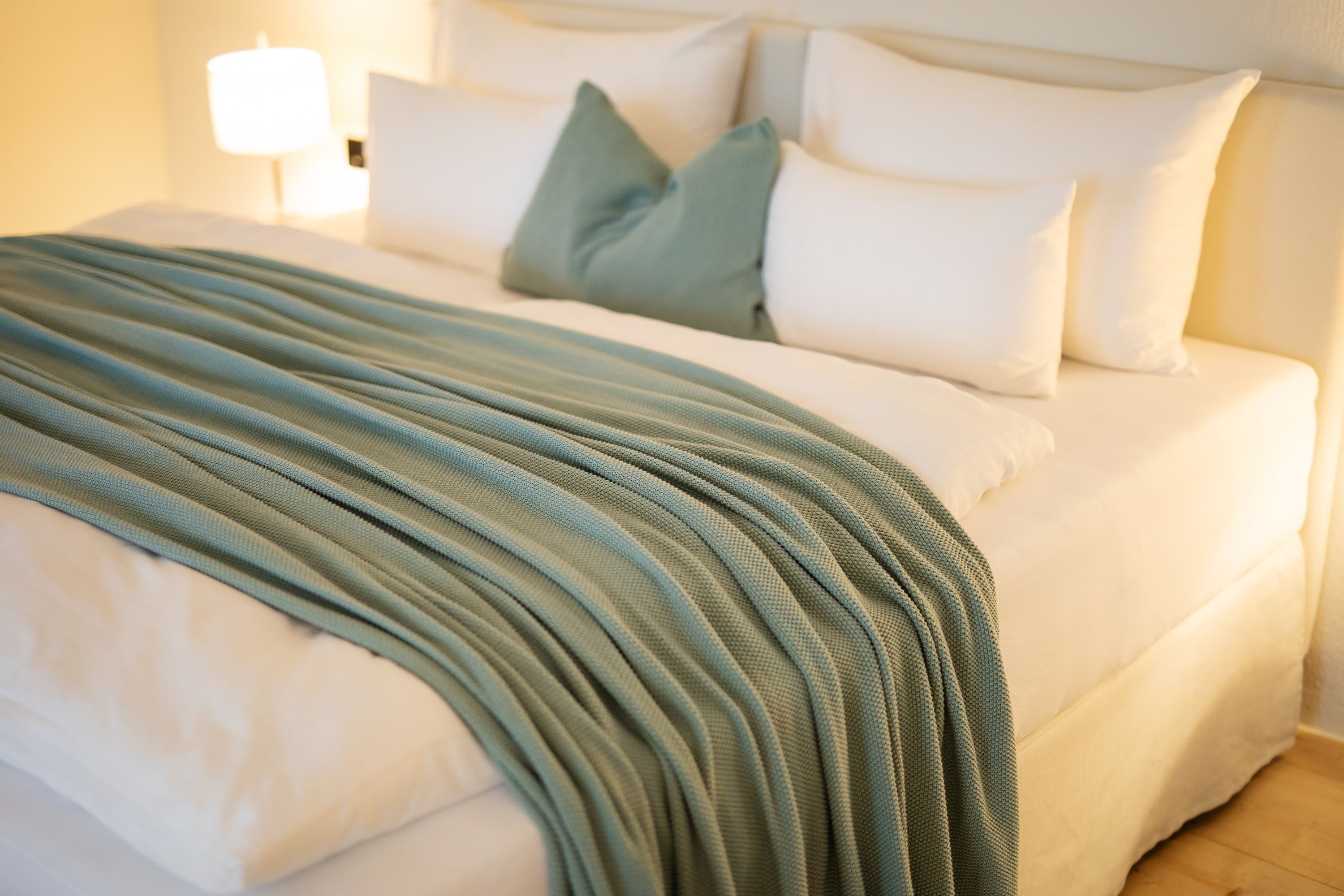 Una cama bien hecha con una manta verde y blanca a rayas, almohadas blancas y una lámpara de noche.
