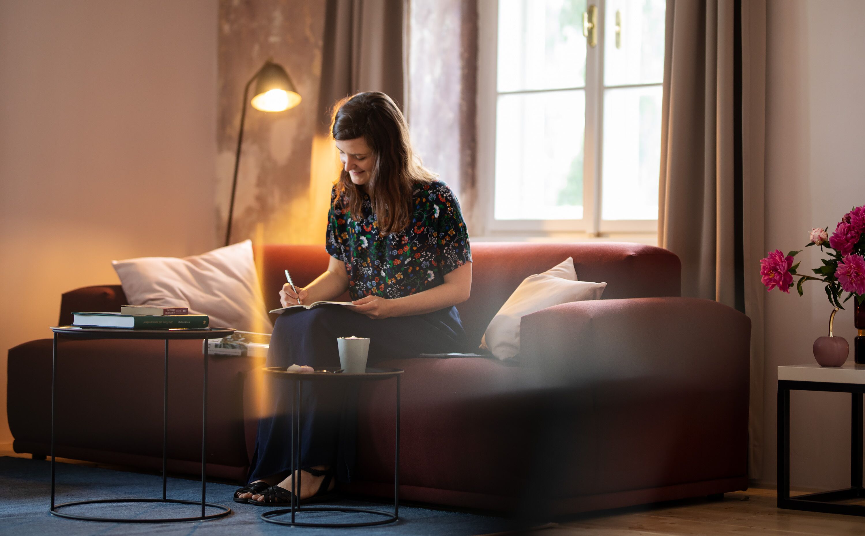 A woman sitting on a red sofa, writing in a notebook. There is a lamp, a small table with a book, and a vase with flowers on a side table.