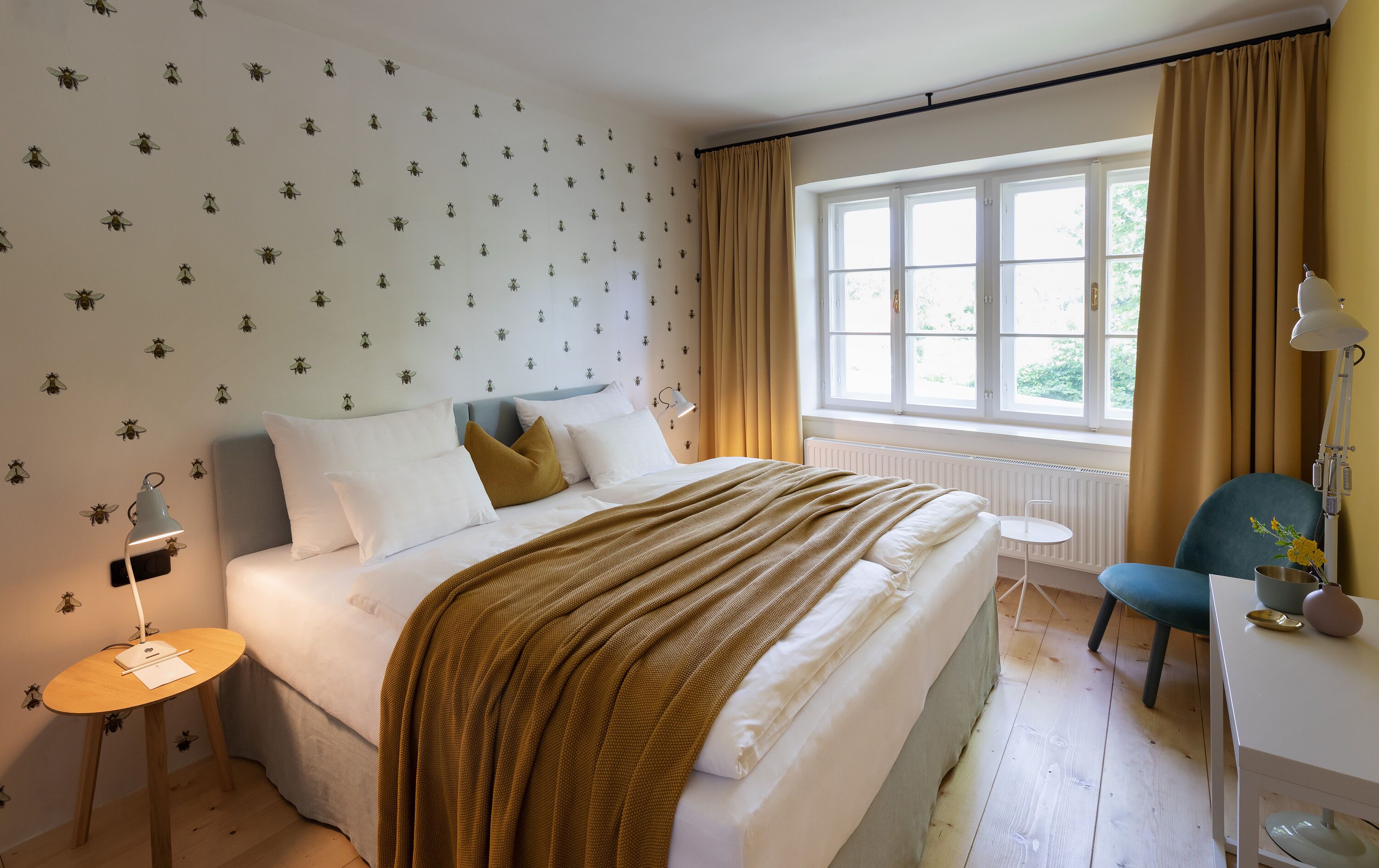 Een slaapkamer met een bed, nachtkastje, lamp, stoel en raam met gordijnen.