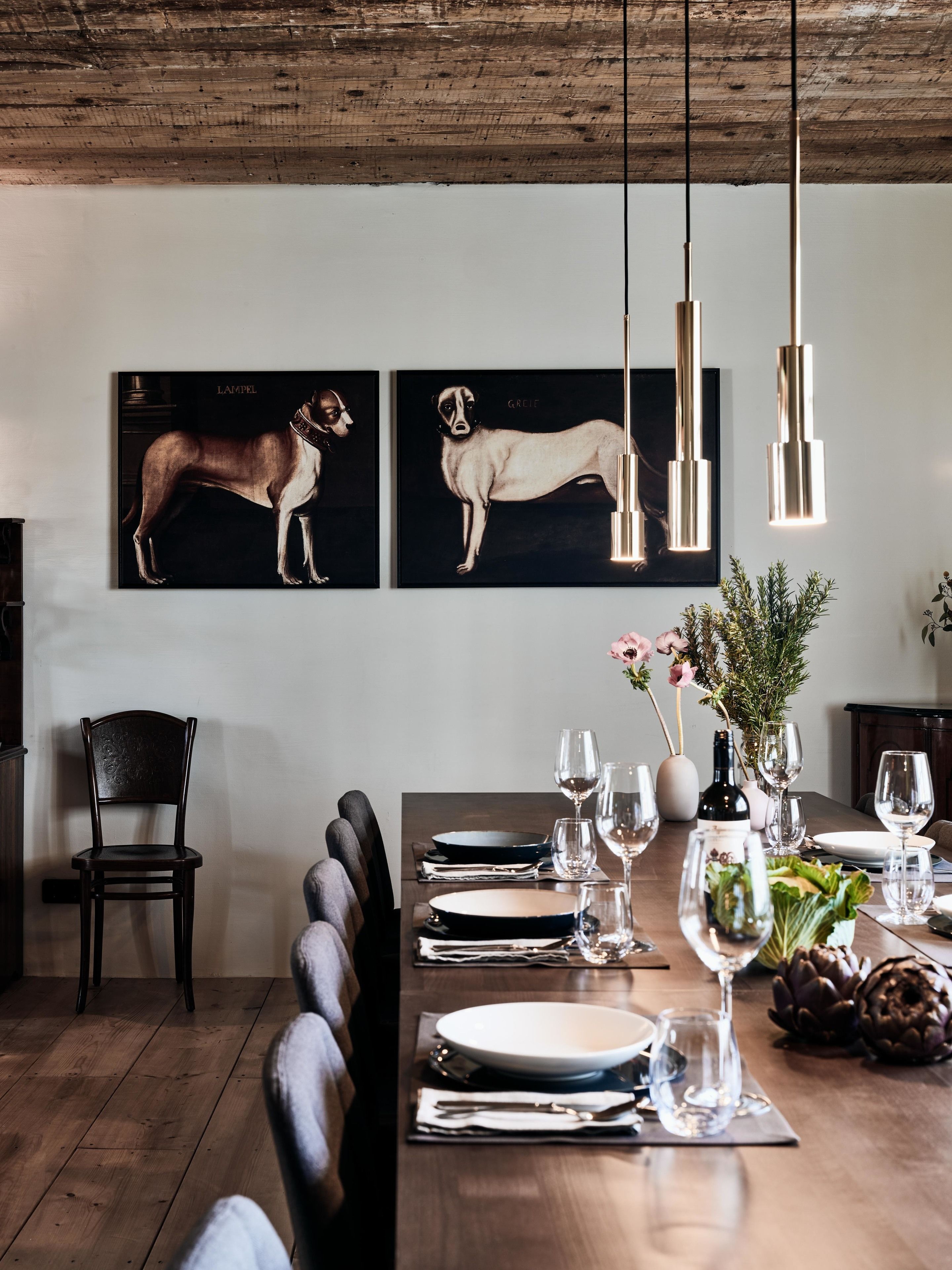 Een eethoek met een houten tafel gedekt voor een maaltijd, twee ingelijste hondenportretten aan de muur en hangende pendantlampen.