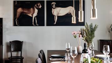 Een eethoek met een houten tafel gedekt voor een maaltijd, twee ingelijste hondenportretten aan de muur en hangende pendantlampen.