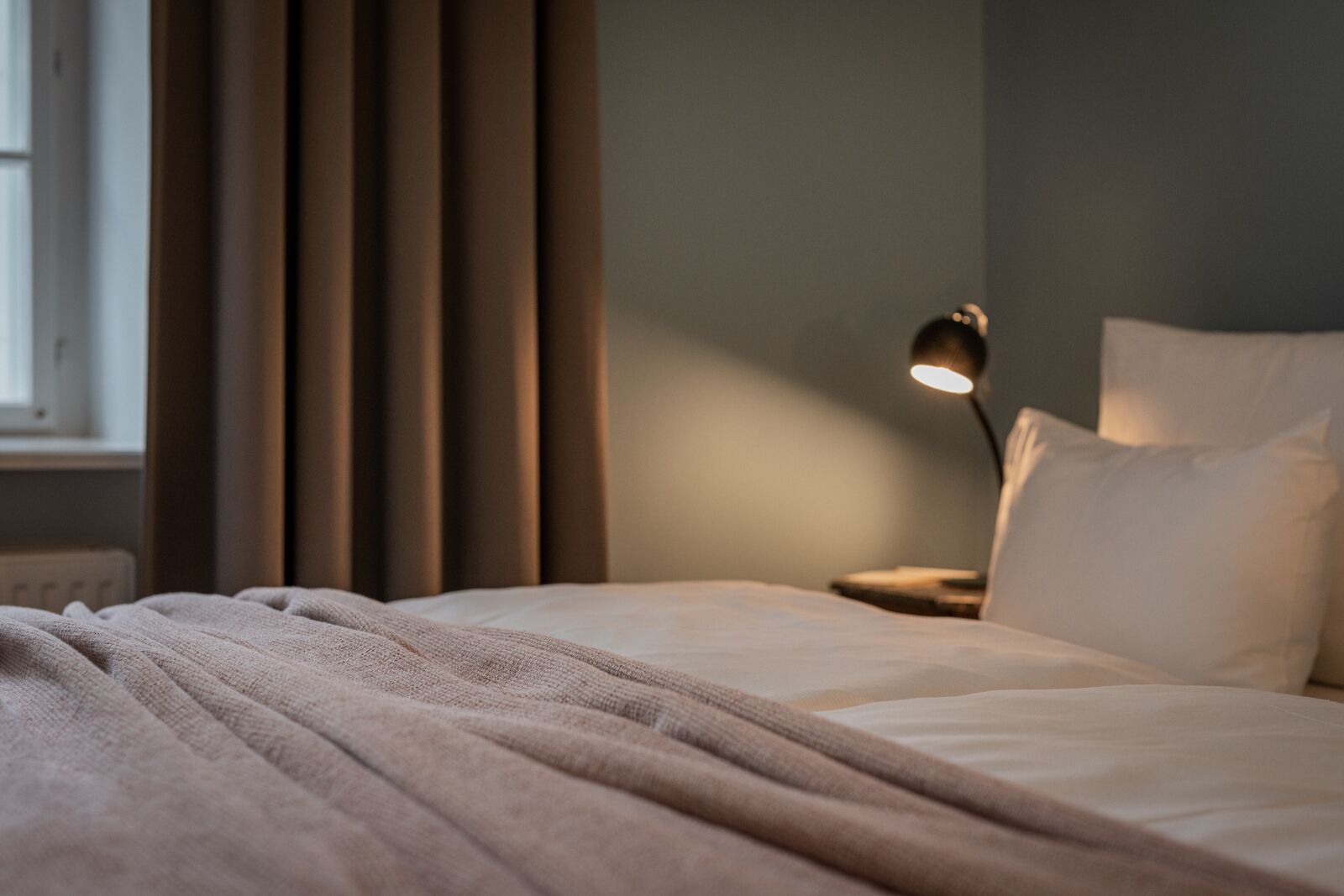 Een net opgemaakt bed met een deken, een bedlampje en een raam met gordijnen.