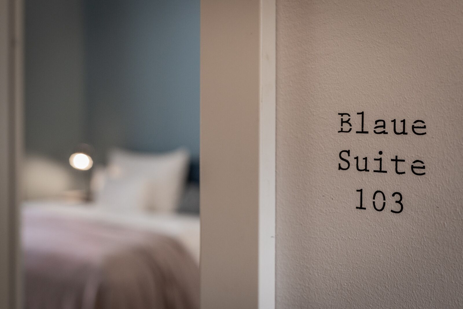 A room with a bed and a nightstand, a wall with the text "Blau Suite 103".