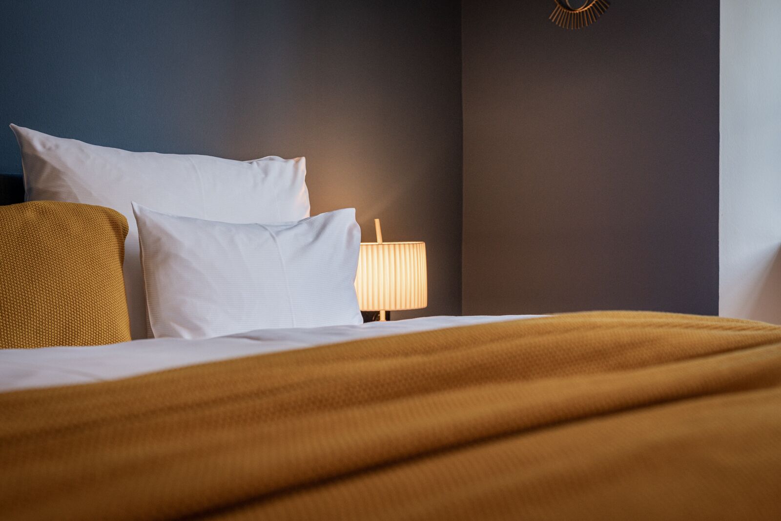 A neatly made bed with white and mustard-colored bedding, a bedside lamp, and a dark wall with a gold decorative element.