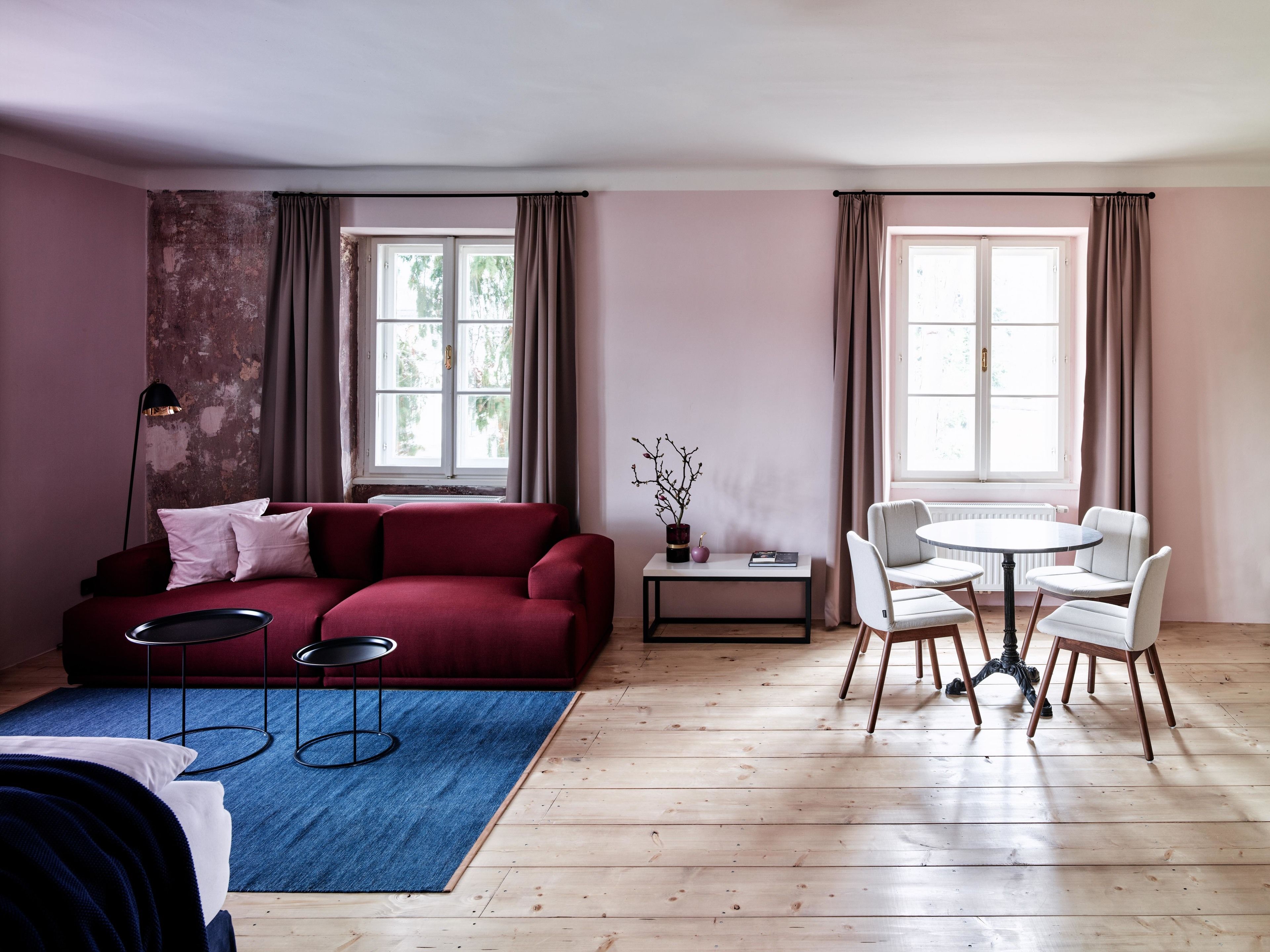 Un salon moderne avec un canapé rouge, un tapis bleu, une table ronde entourée de quatre chaises et deux fenêtres avec des rideaux.