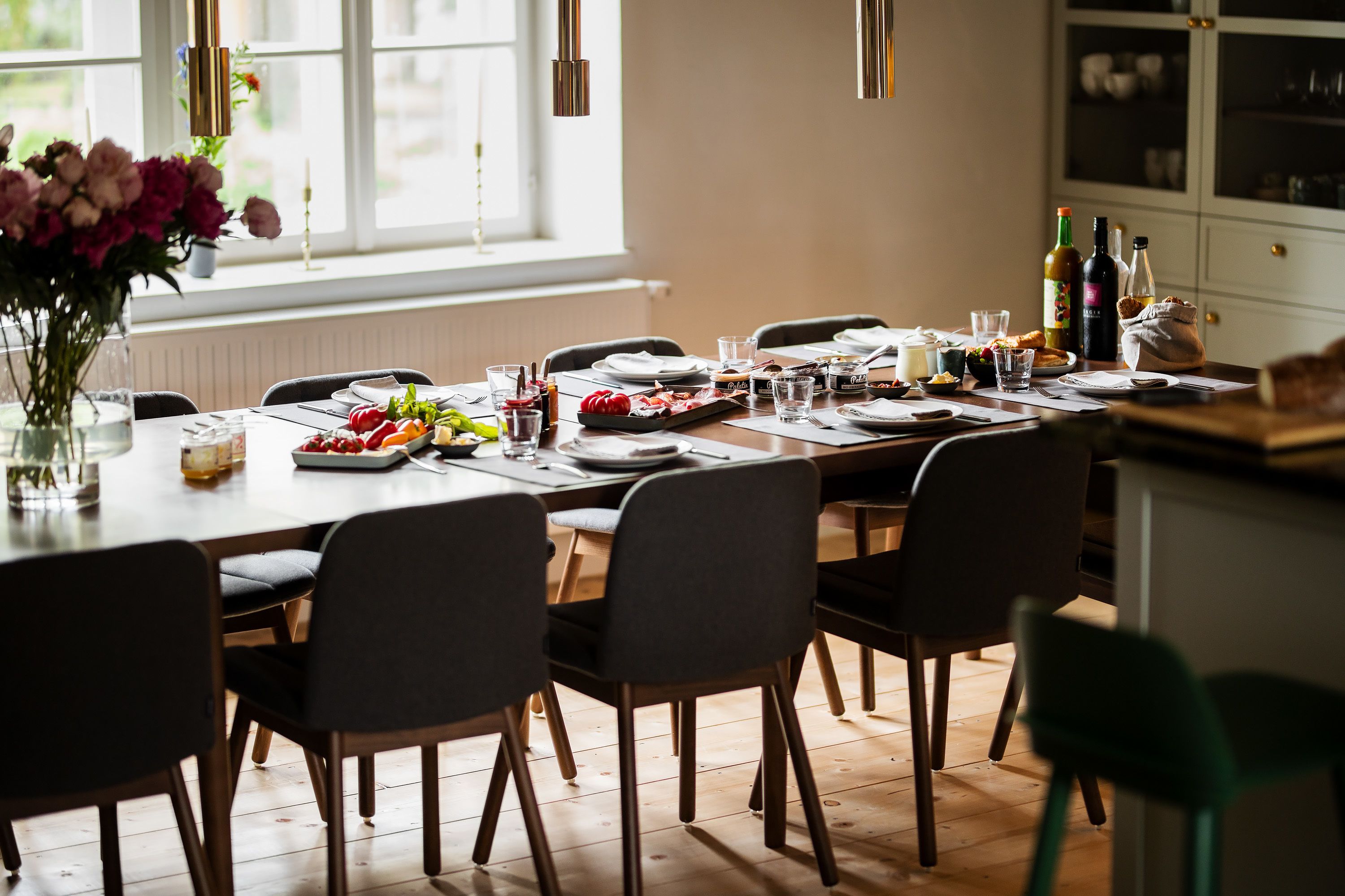 A dining area with a wooden table set for a meal, chairs, a vase with flowers, and a window with natural light.