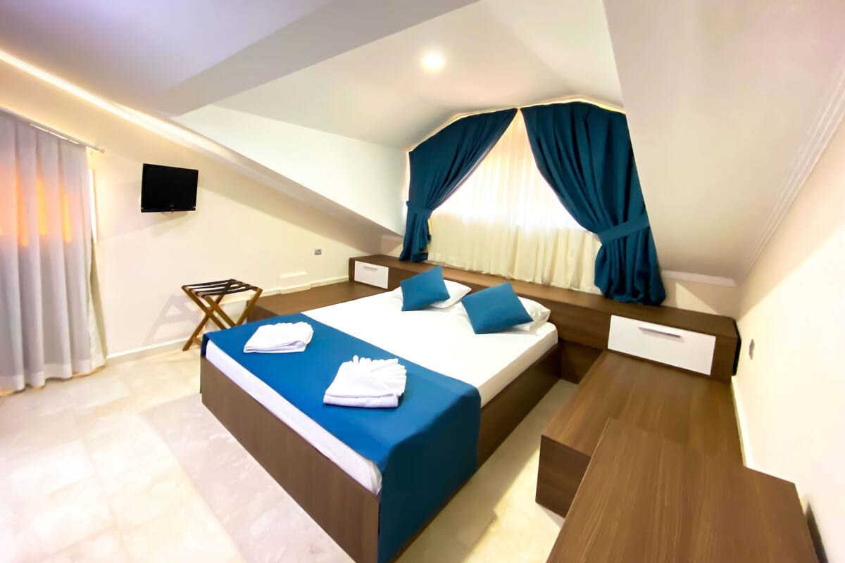 Une chambre d’hôtel moderne dotée d’un grand lit, d’une table de chevet en bois, d’un téléviseur à écran plat et d’une fenêtre avec des rideaux.
