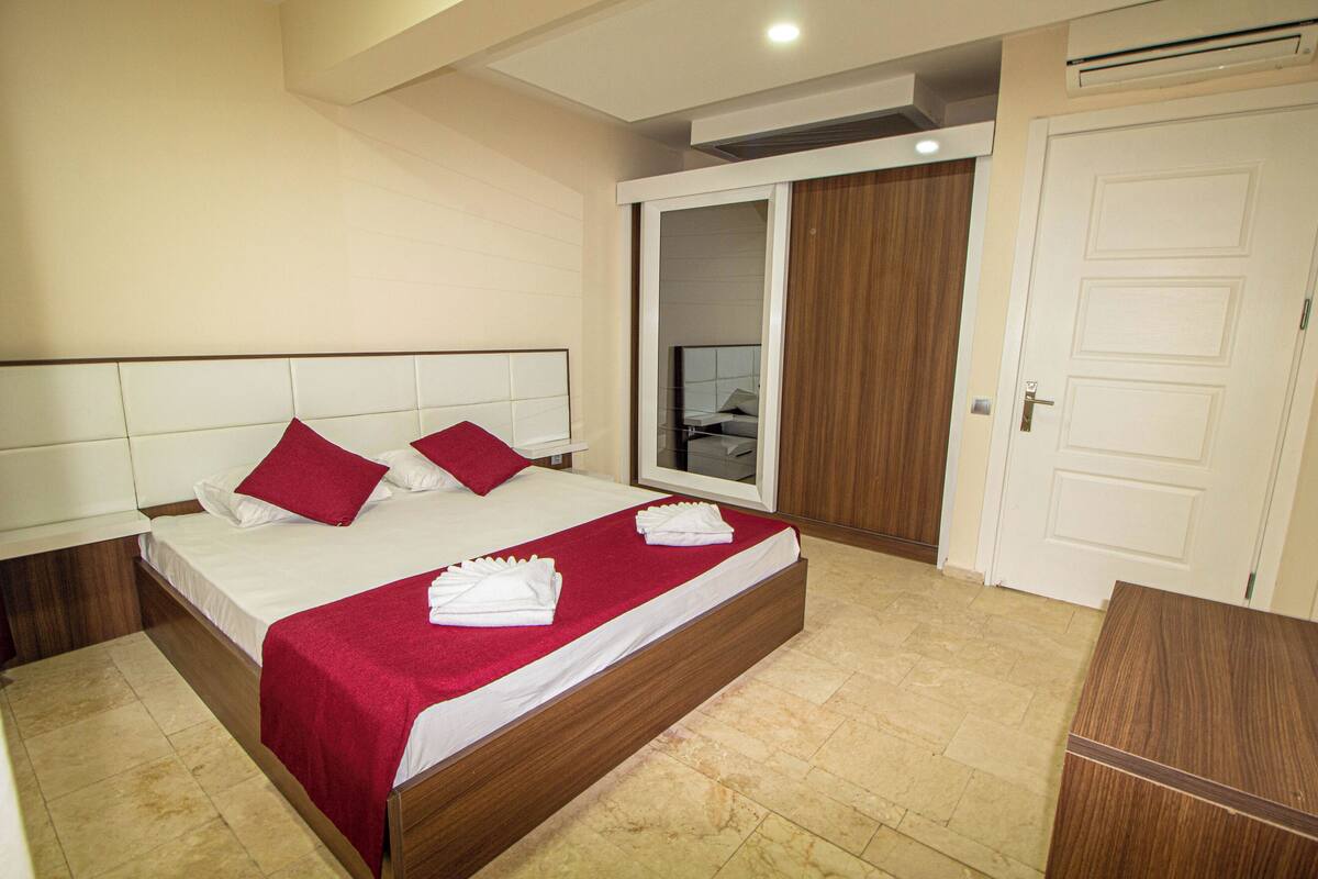 Une chambre d’hôtel avec un grand lit, une tête de lit en bois, une couvre-lit rouge et un miroir au mur.